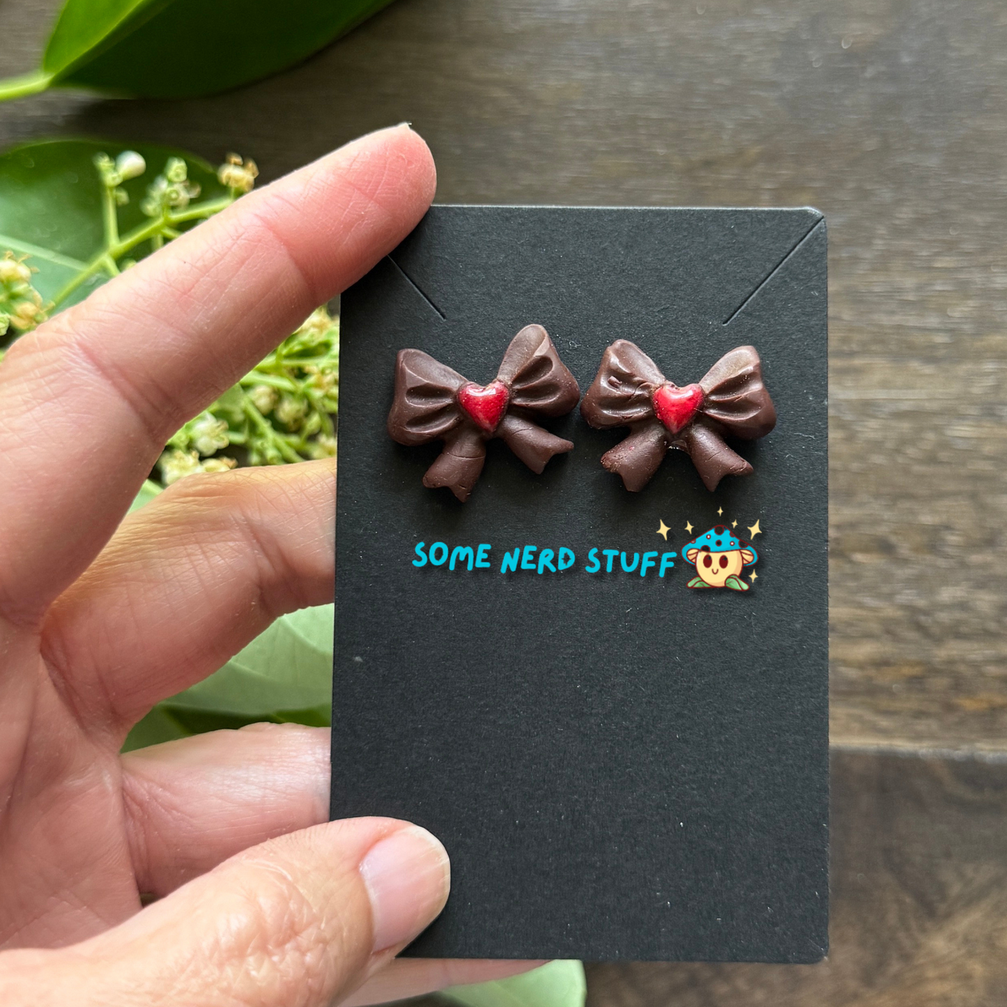 Chocolate bows with red heart center polymer clay handmade earrings held by hand to show size by SomeNerdStuff