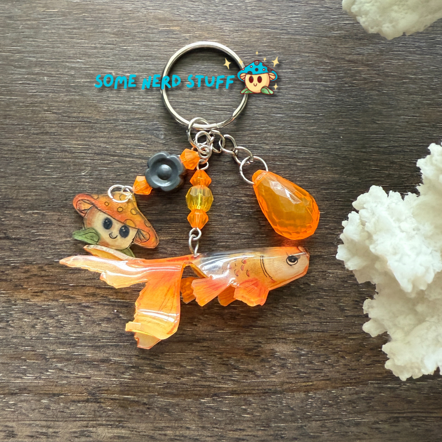 Keychain with orange fish charm, small character charms, and 'Some Weird Stuff' text on a wooden surface.