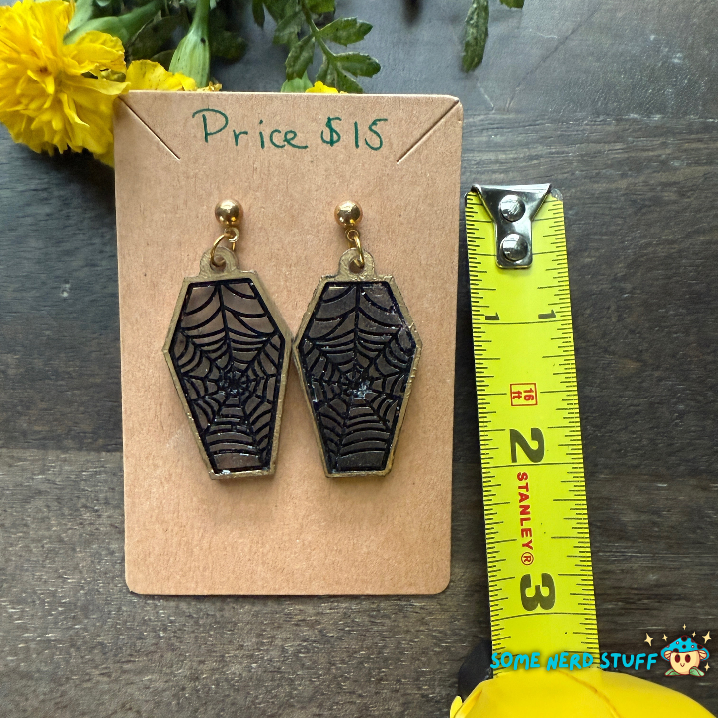 Web coffin-shaped earrings on a card with a measuring tape for scale, on a dark surface sold on somenerdstuff.com