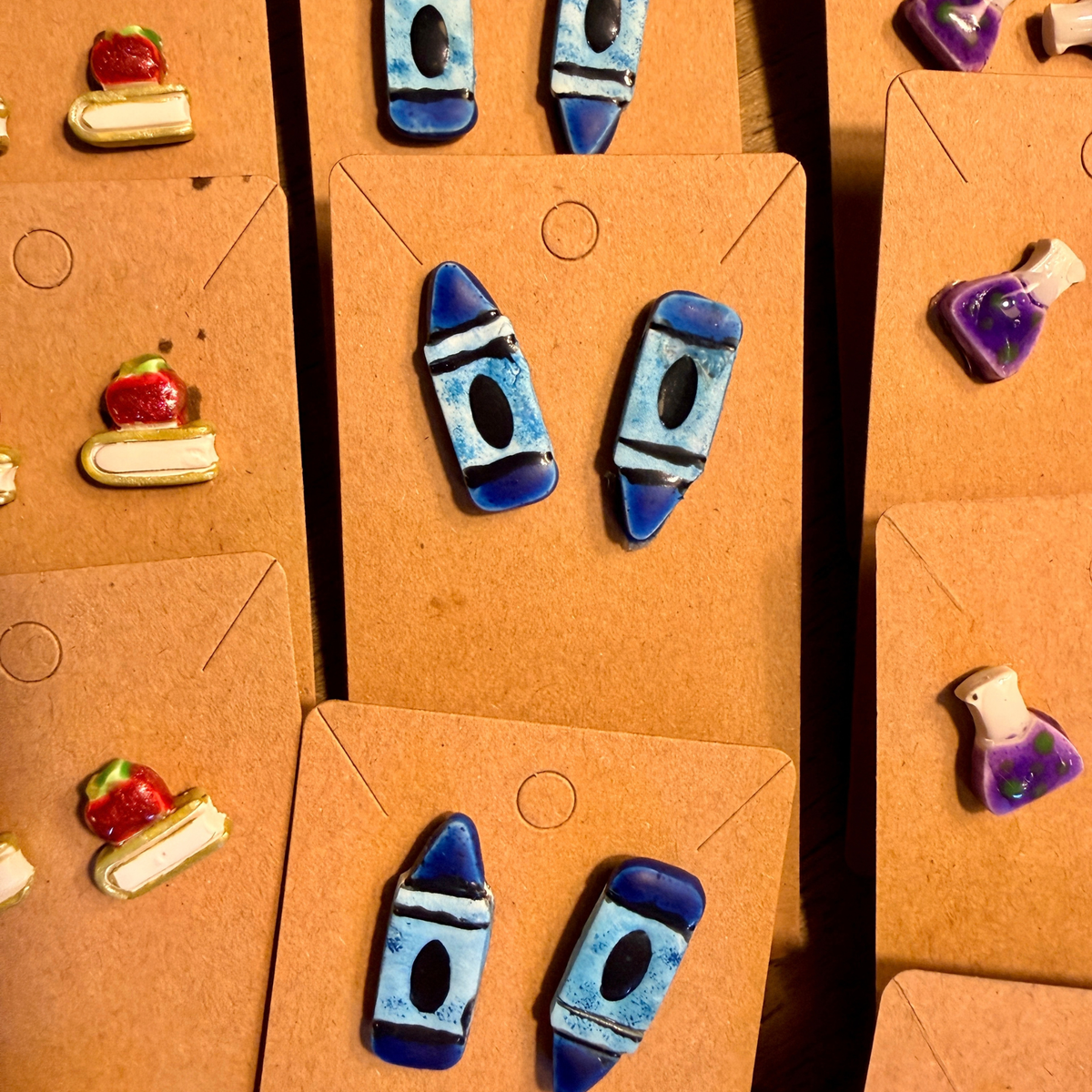 Several pairs of blue crayon stud earrings displayed on kraft cards beside other school-themed earrings.