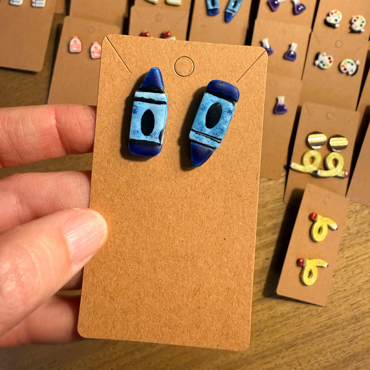 Hand holding blue crayon stud earrings on a kraft card to show their size in context.