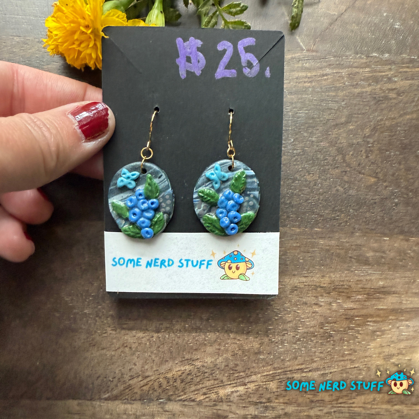Pair of polymer clay handmade earring dangles with faux blueberry branches, blue butterflies, and faux stone with a price tag on a wooden surface.