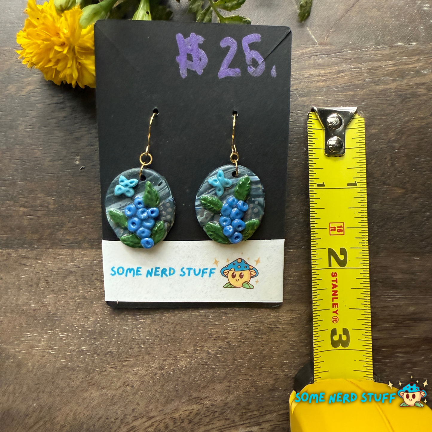 Pair of polymer clay handmade earring dangles with faux blueberry branches, blue butterflies, and faux stone with a price tag on a wooden surface showing size