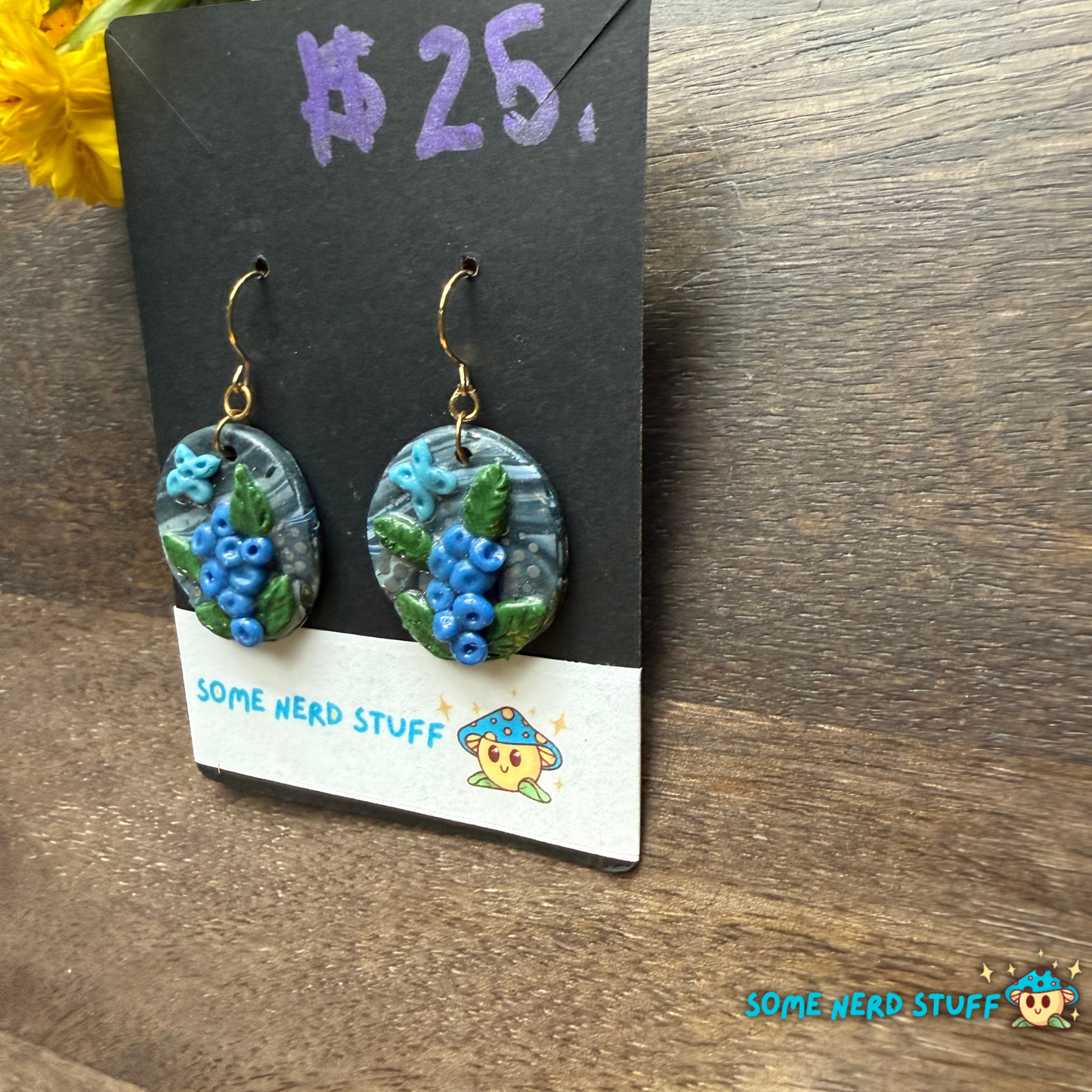 Pair of polymer clay handmade earring dangles with faux blueberry branches, blue butterflies, and faux stone with a price tag on a wooden surface.