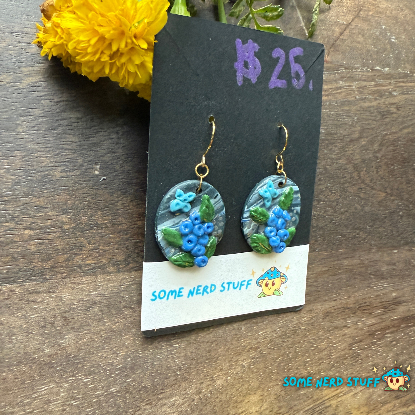 Pair of polymer clay handmade earring dangles with faux blueberry branches, blue butterflies, and faux stone with a price tag on a wooden surface.