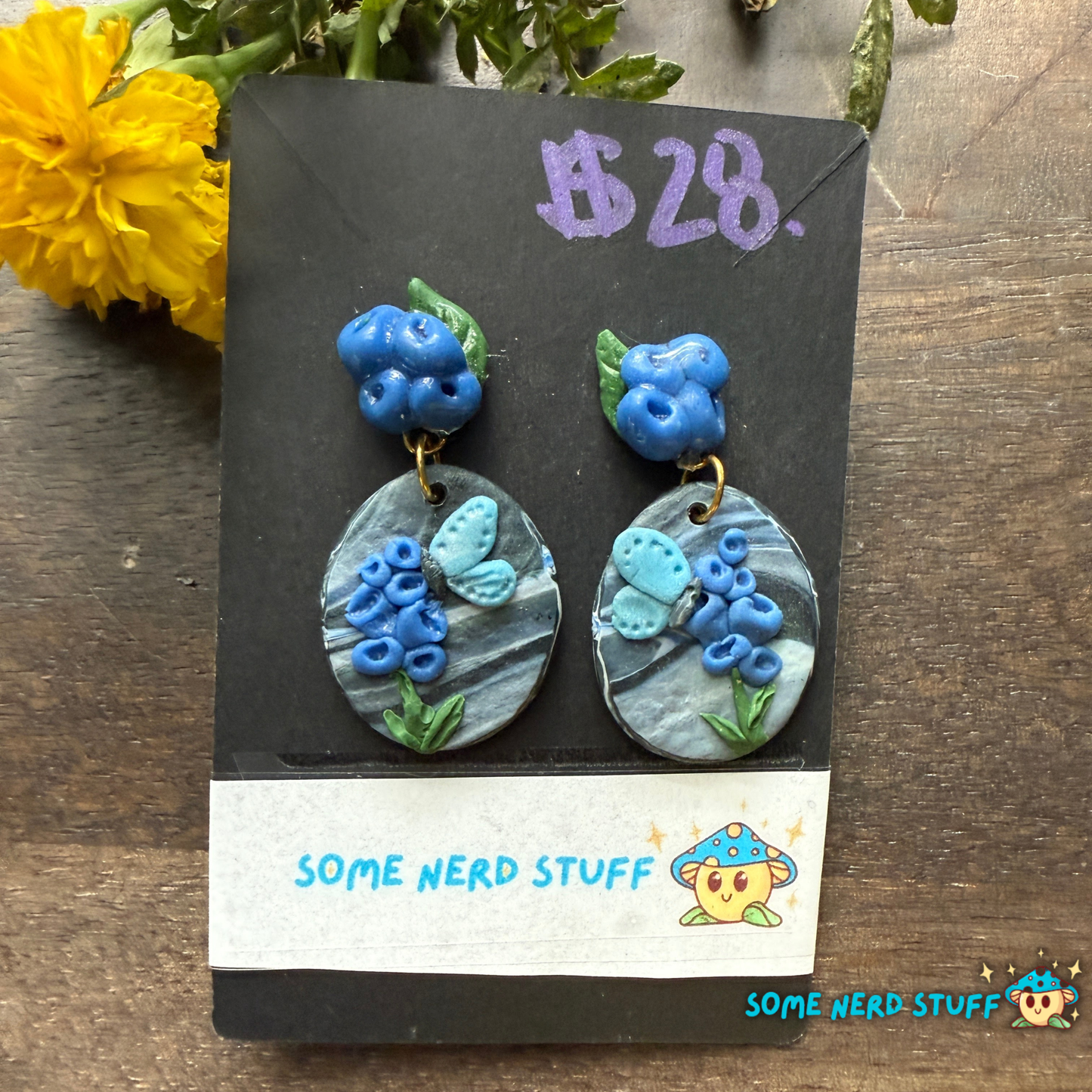 Pair of blue floral earrings on a 'Some Nerd Stuff' branded card with flowers in the background.