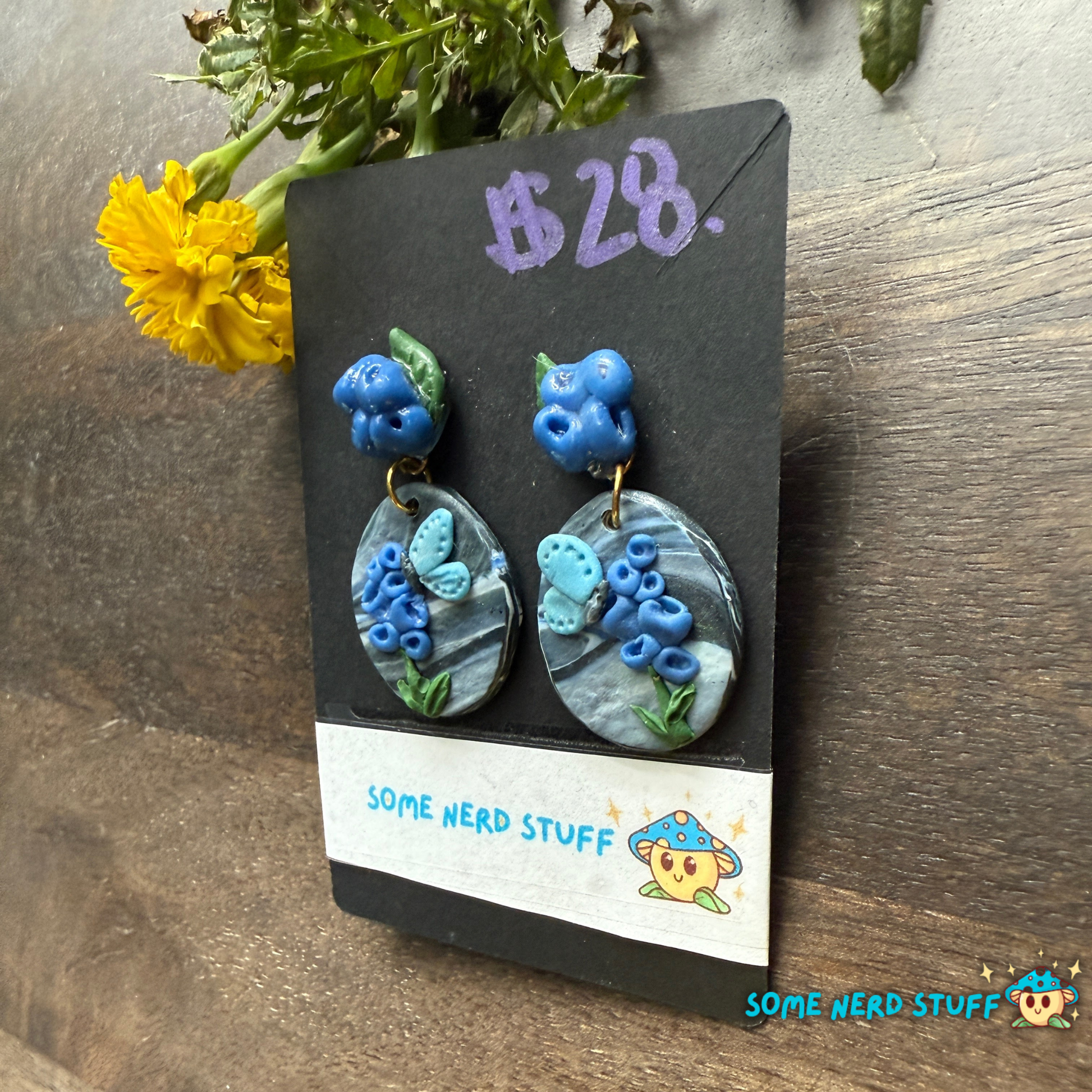 Pair of blueberry-themed earrings on a 'Some Nerd Stuff' branded card with a yellow flower in the background.