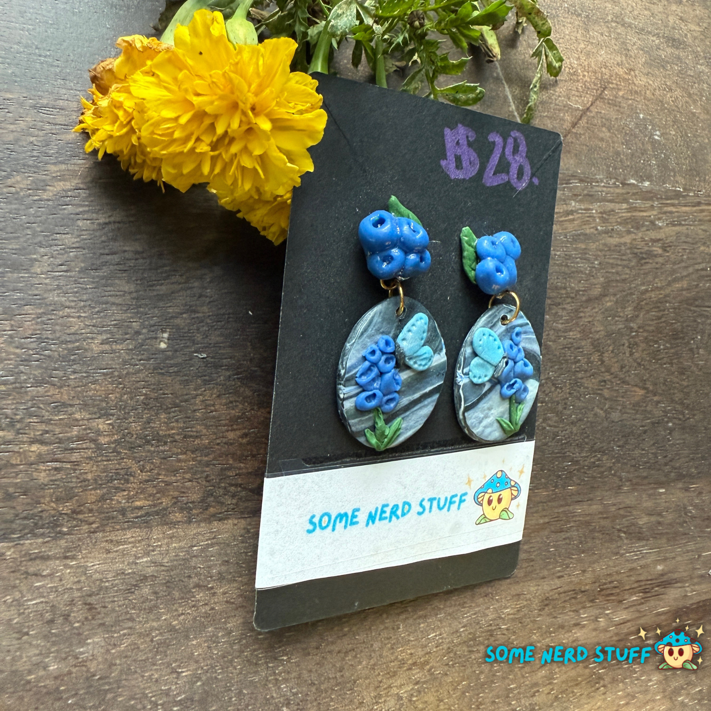 Blue floral earrings on a 'Some Nerd Stuff' branded card with yellow flowers in the background.