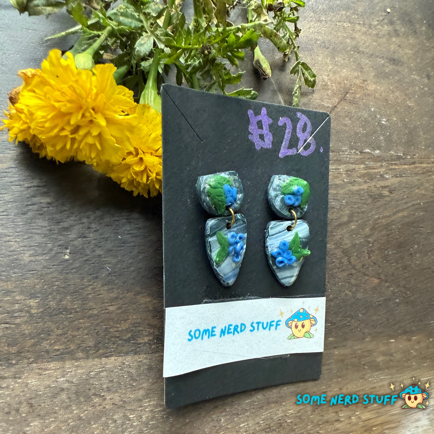 Pair of blueberry and faux stone handmade polymer clay earrings on a 'Some Nerd Stuff' branded card with flowers in the background.