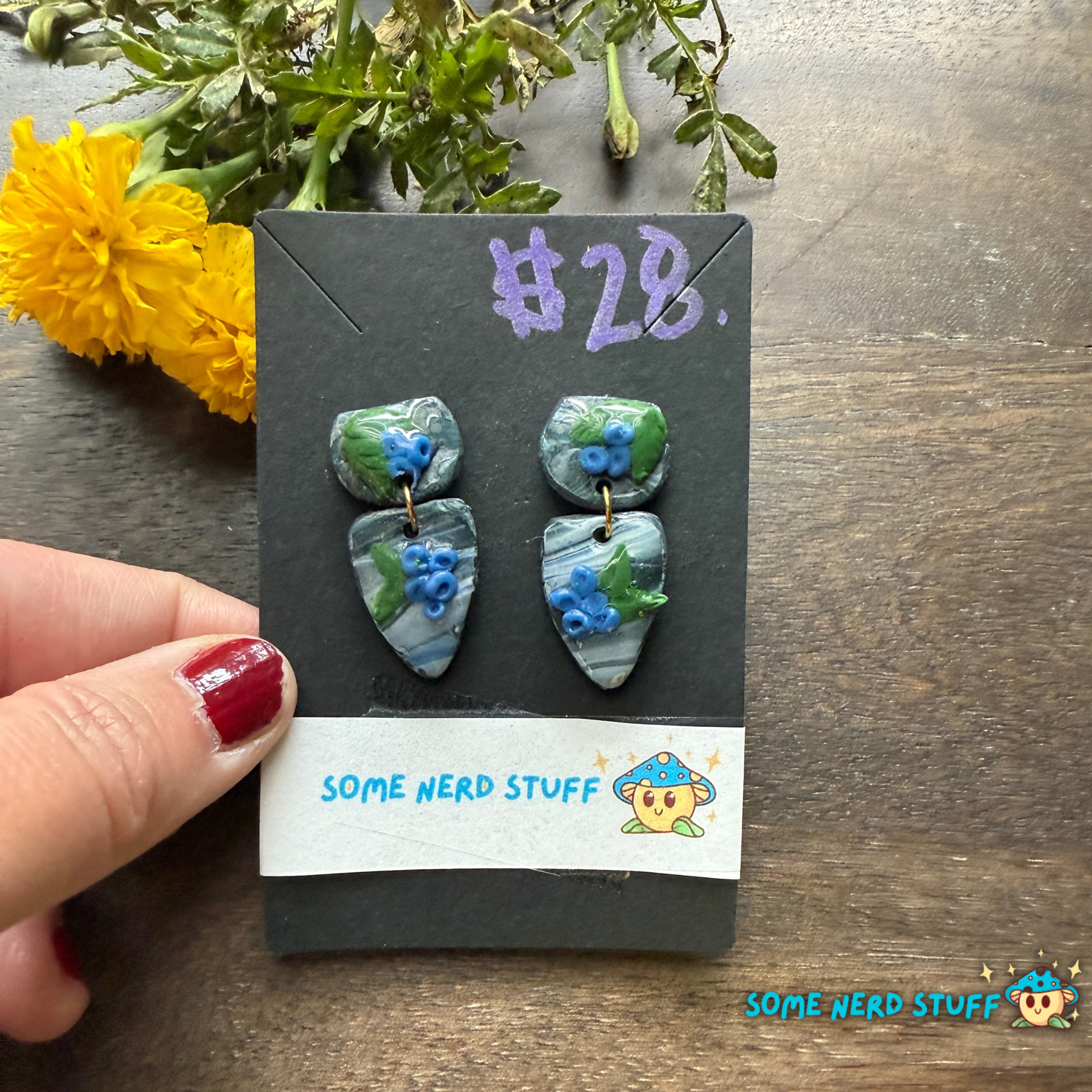 Pair of blueberry and faux stone handmade polymer clay earrings on a 'Some Nerd Stuff' branded card with flowers in the background.