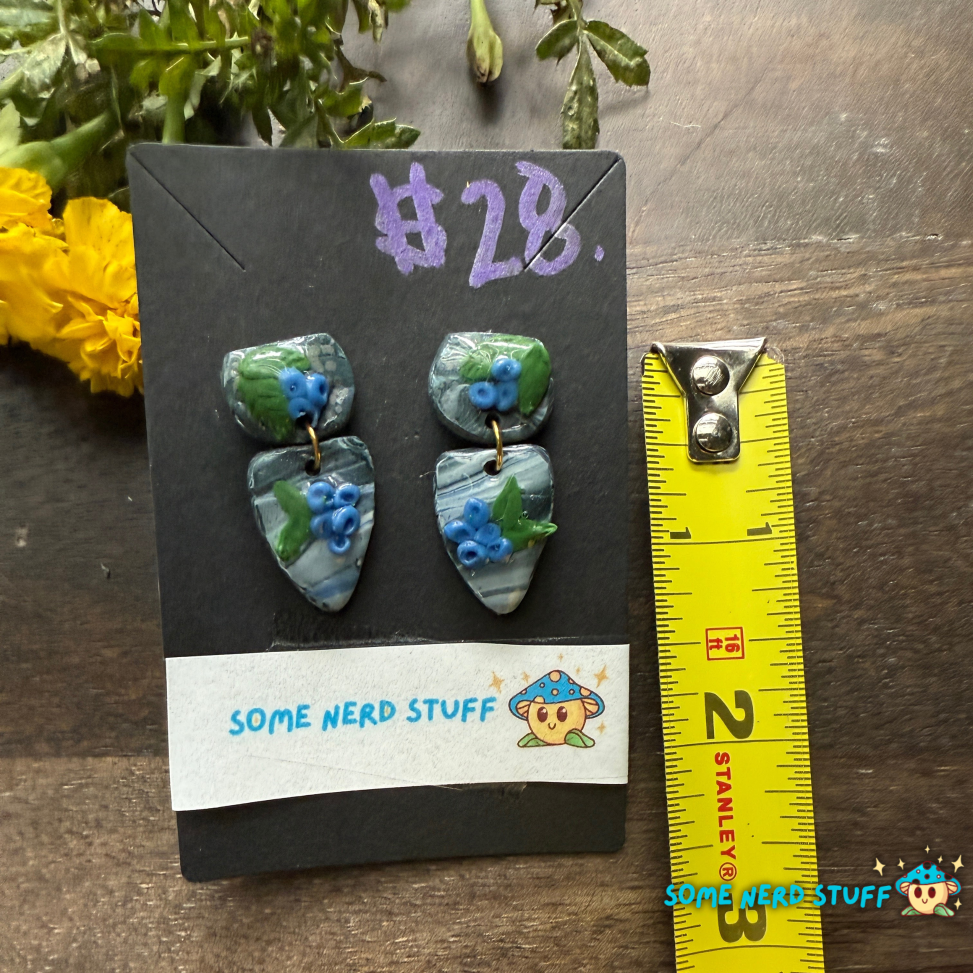 Pair of blueberry and faux stone handmade polymer clay earrings on a 'Some Nerd Stuff' branded card with flowers in the background showing size.