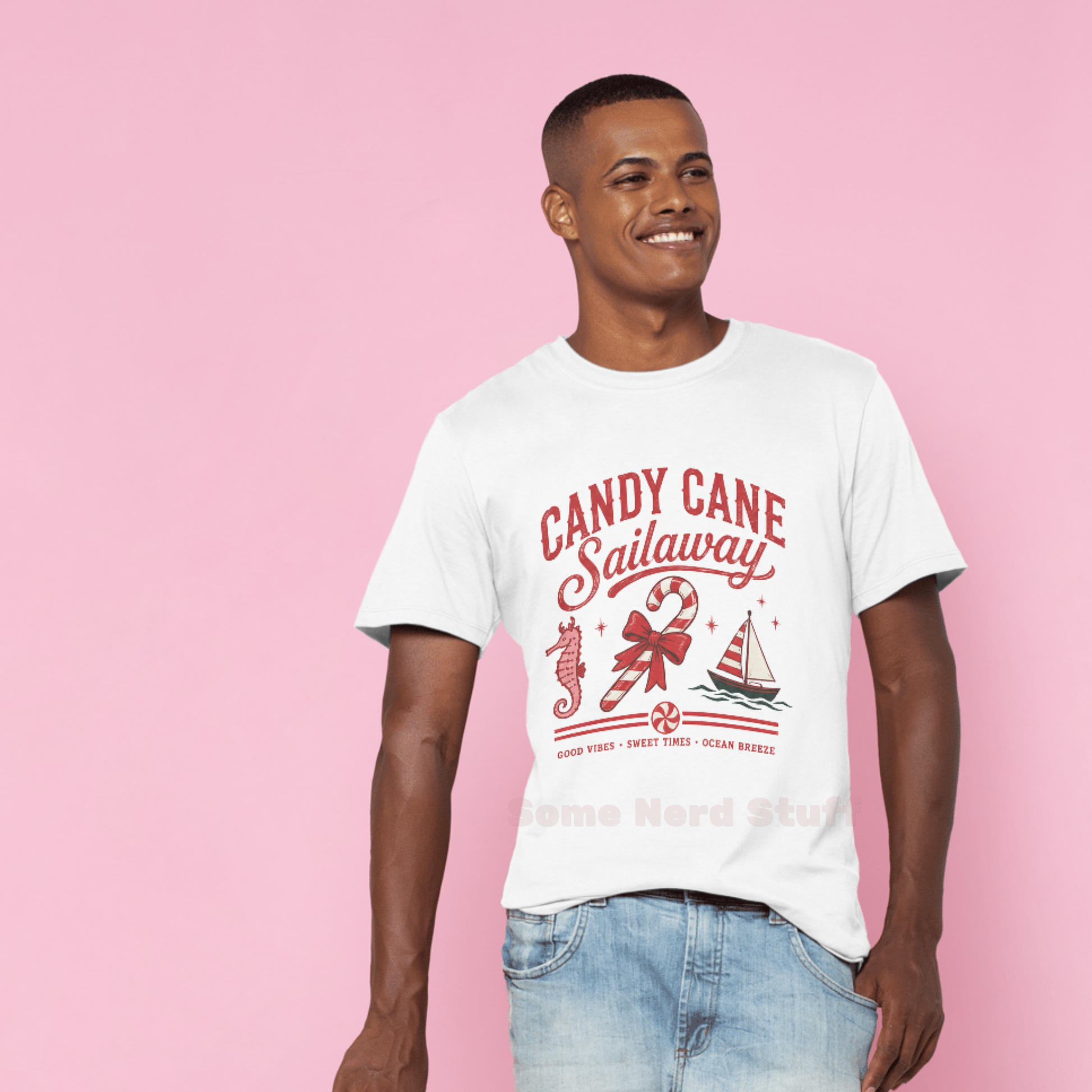 A man wearing a white graphic t-shirt with a red 'Candy Cane Sailaway' design, paired with light blue jeans, standing against a solid pink background.