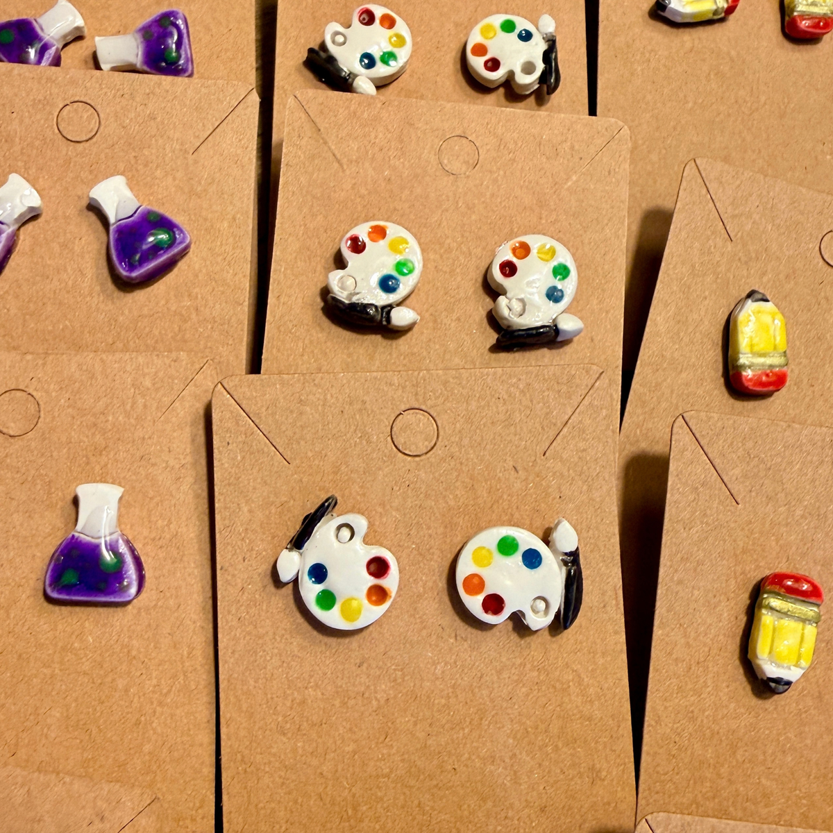 Several pairs of white paint palette stud earrings with colorful paint dots displayed on kraft cards.