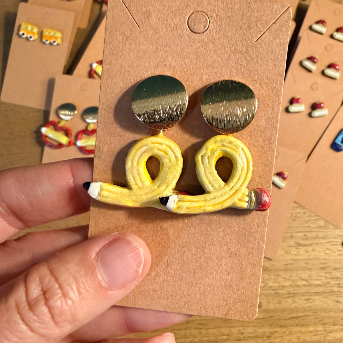 Hand holding looped yellow pencil dangle earrings with round gold-tone tops on a kraft card.