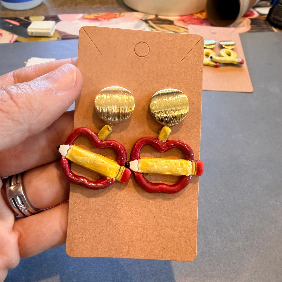 Hand holding a pair of apple and pencil dangle earrings with round gold-tone tops on a kraft card.