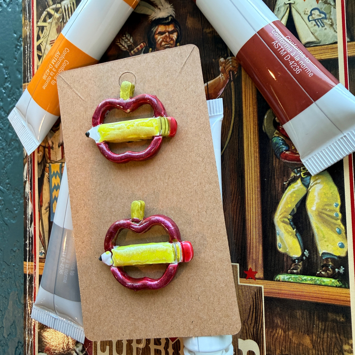 Pair of red apple stud earrings with yellow pencil details on a kraft display card.