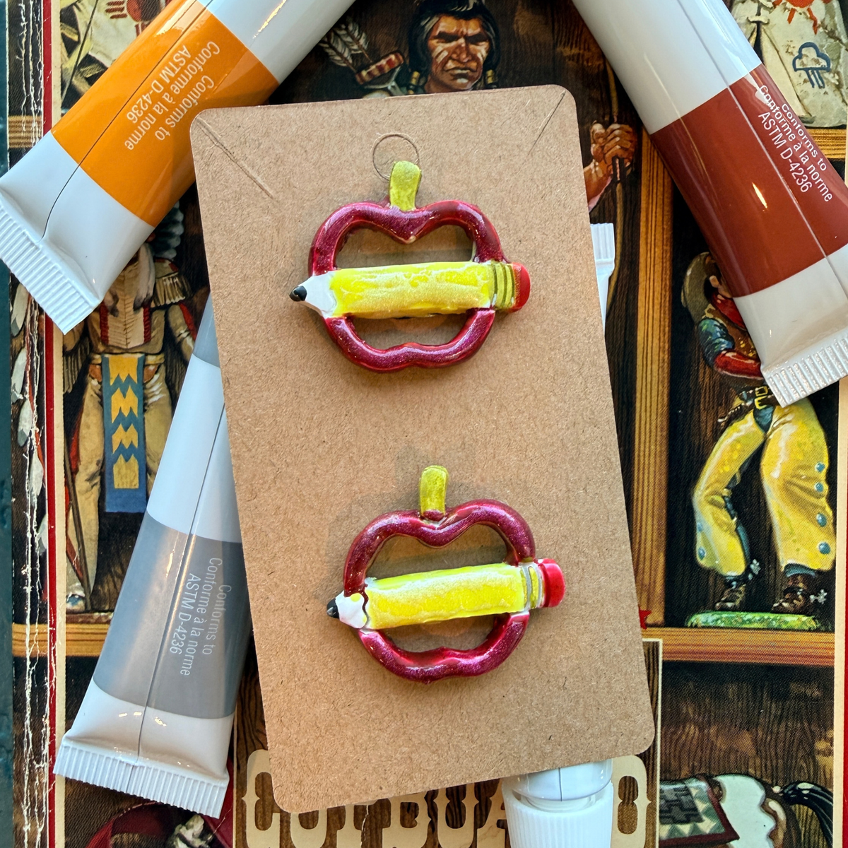 Front view of matching apple and pencil stud earrings on a display card with art supplies behind them.