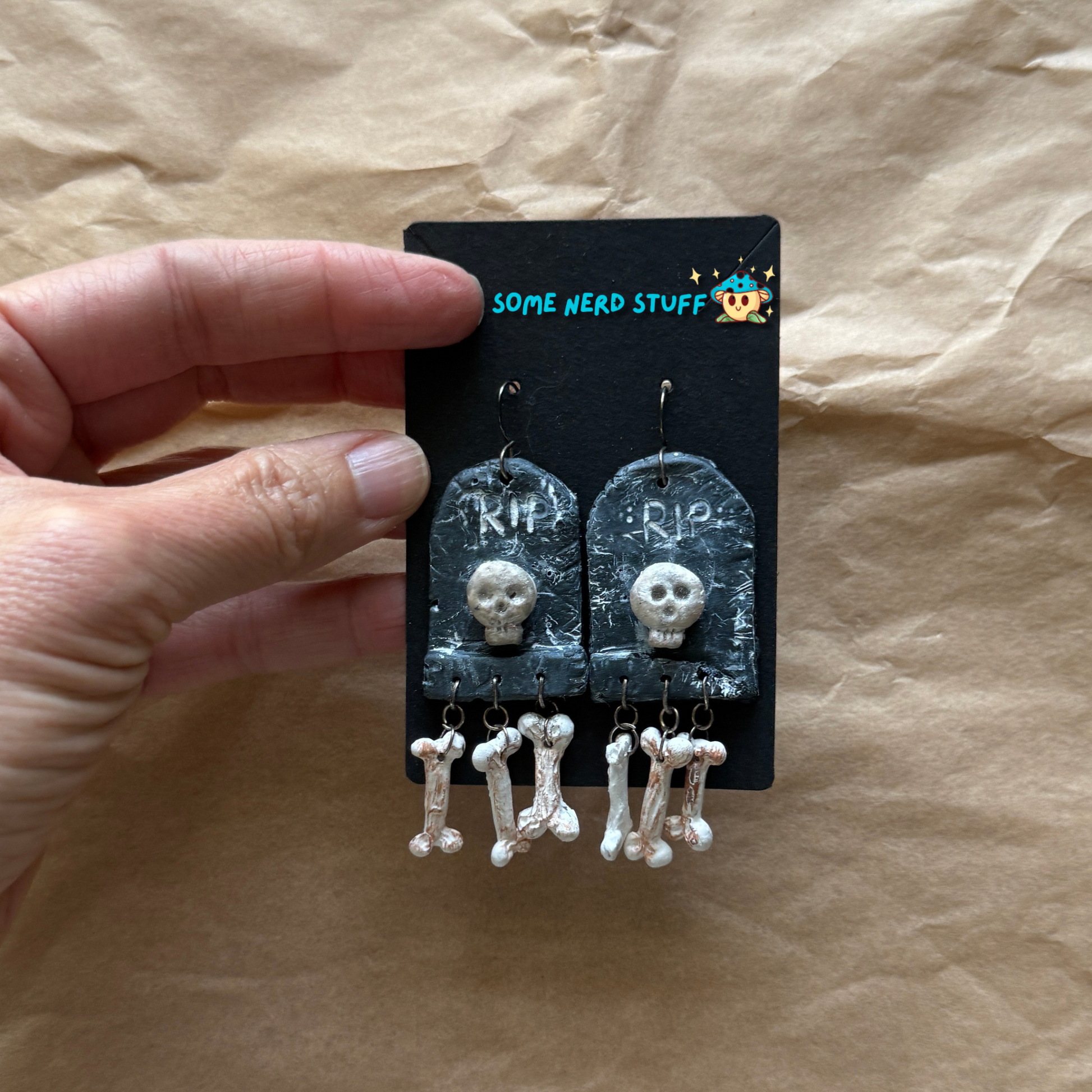 Phntom Parlor Polymer Clay tombstone skull and dangling bone earrings held by hand to show size made by SomeNerdStuff