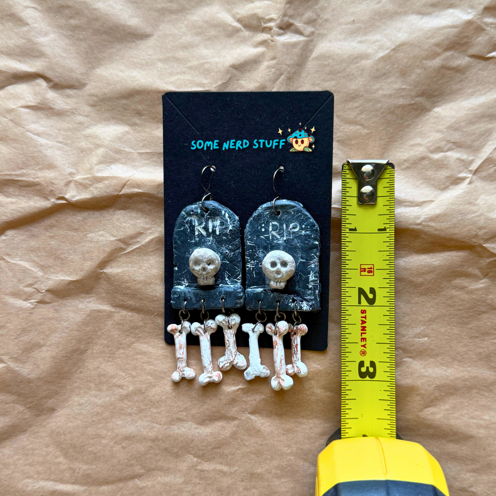 Phntom Parlor Polymer Clay tombstone skull and dangling bone earrings shown with measuring tape made by SomeNerdStuff