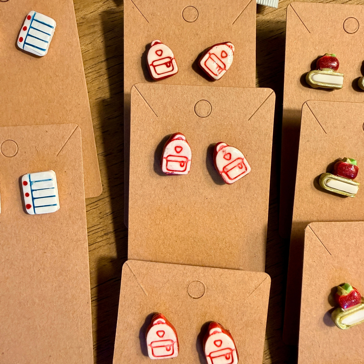Pink and white backpack stud earrings with outlined pockets displayed on kraft cards.