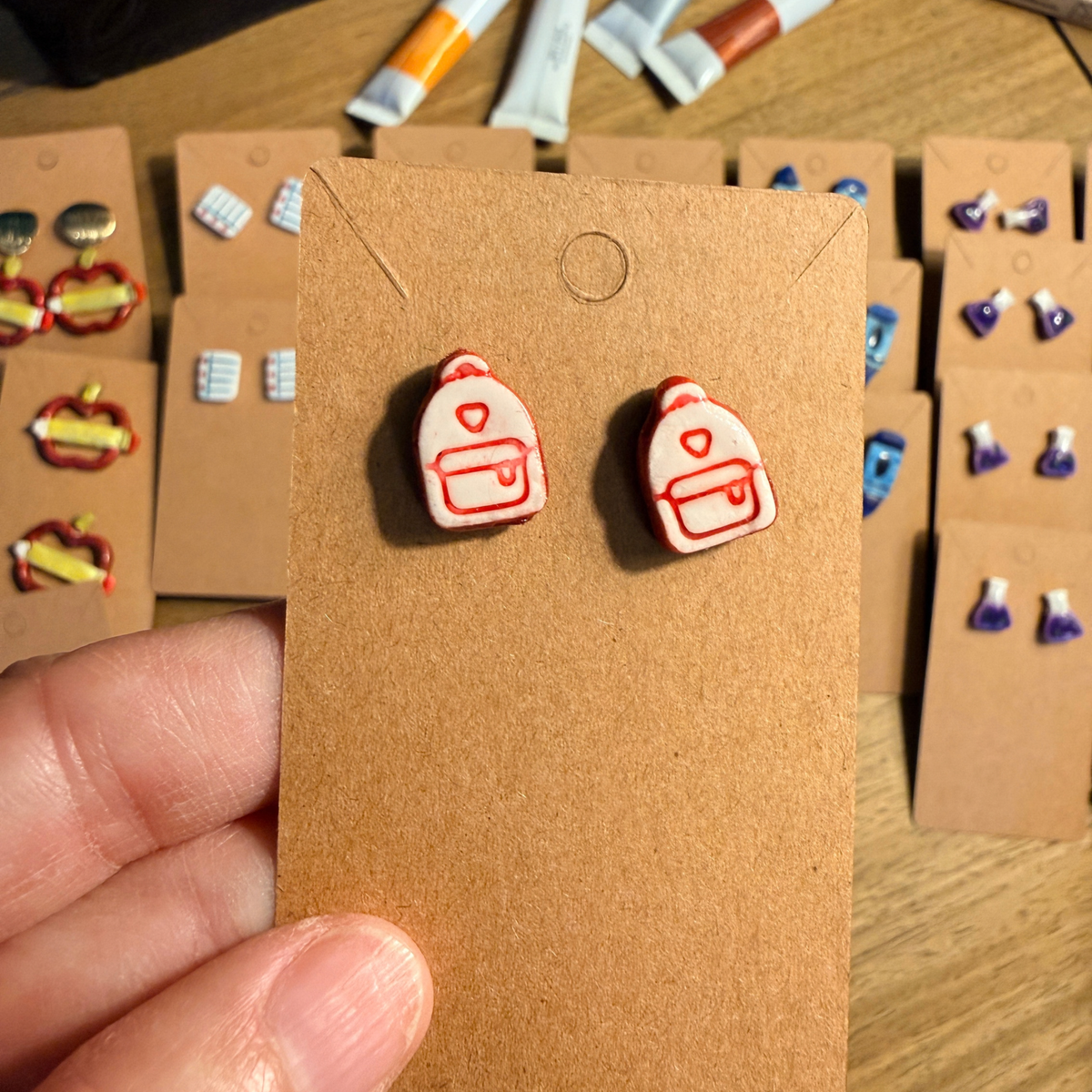 Hand holding a matching pair of pink backpack stud earrings on a kraft display card.