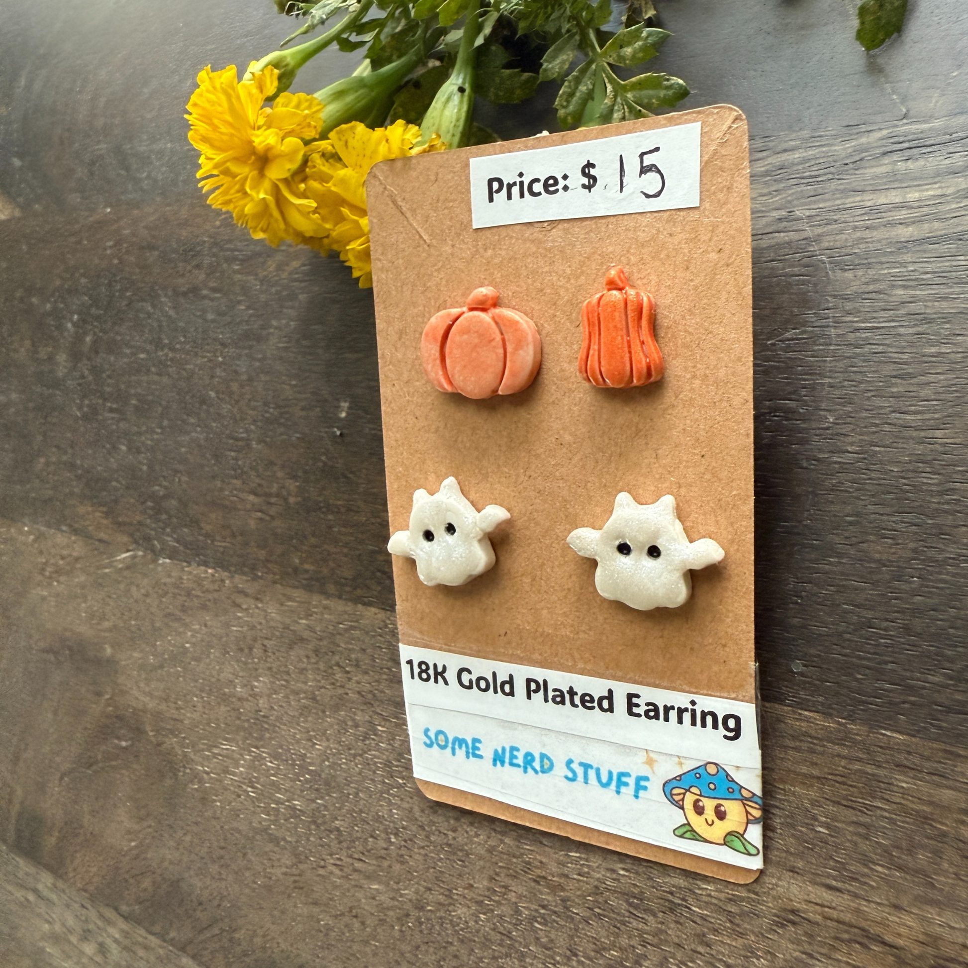 Halloween-themed earrings on a cardboard backing with a price tag, on a wooden surface featuring pumpkins and flying ghosts.