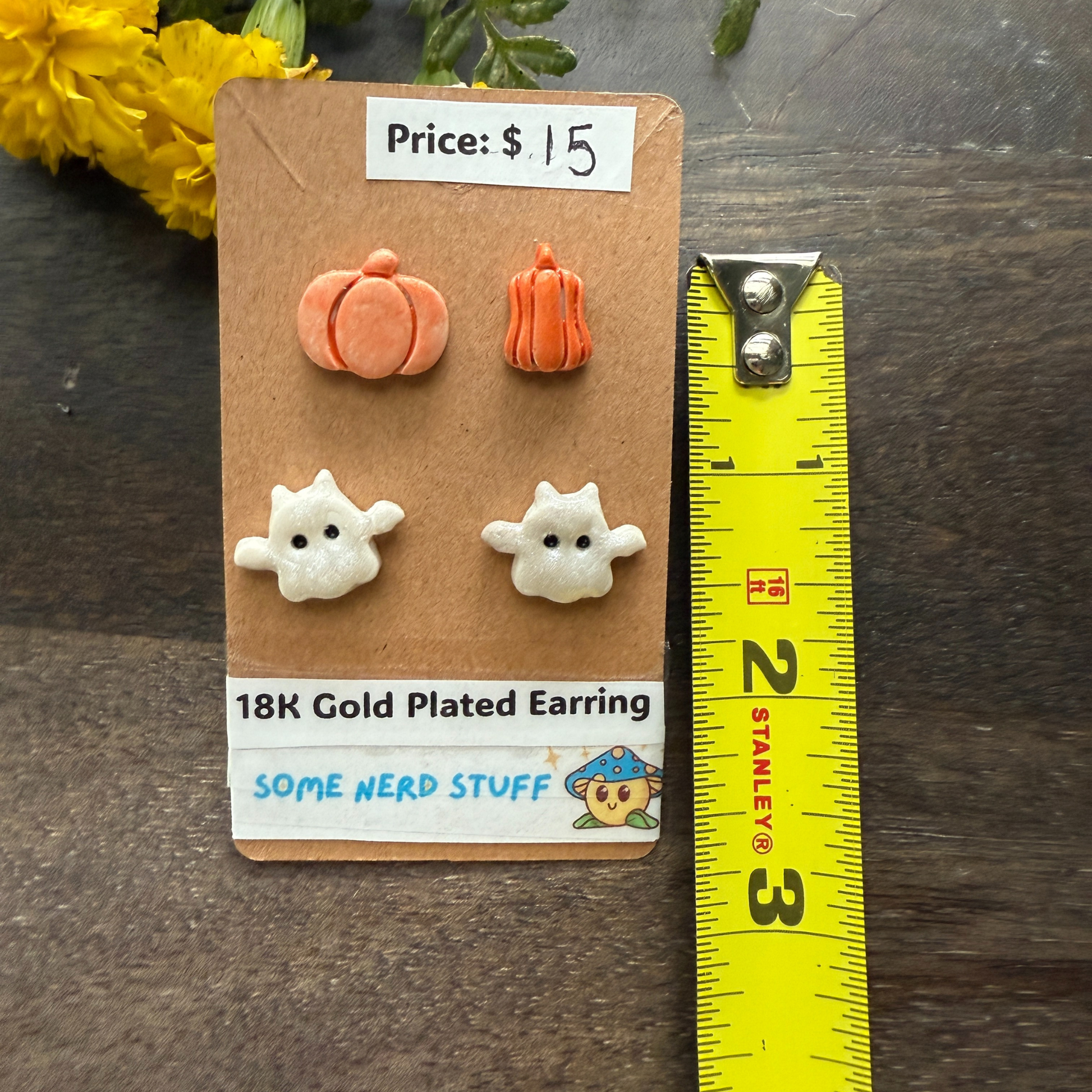 Pair of pumpkin and ghost earrings on a card with a ruler for scale, on a wooden surface.
