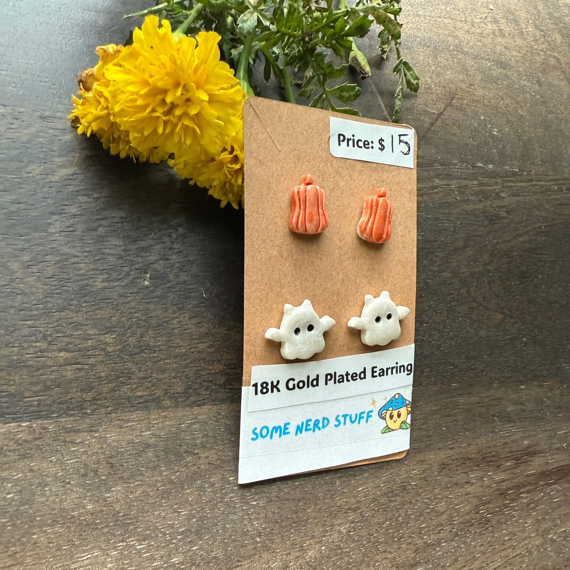 Halloween-themed earrings on a cardboard backing with a price tag, on a wooden surface featuring pumpkins and flying ghosts 18k gold plated earrings.