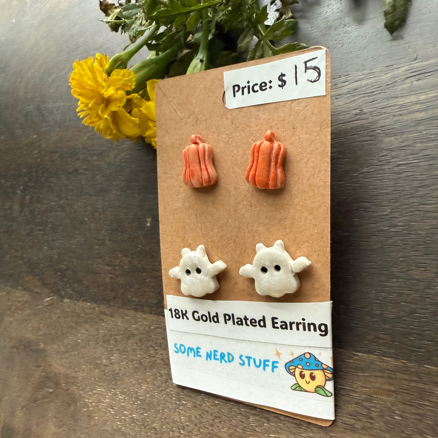 Halloween-themed earrings on a cardboard backing with a price tag, on a wooden surface featuring pumpkins and flying ghosts.