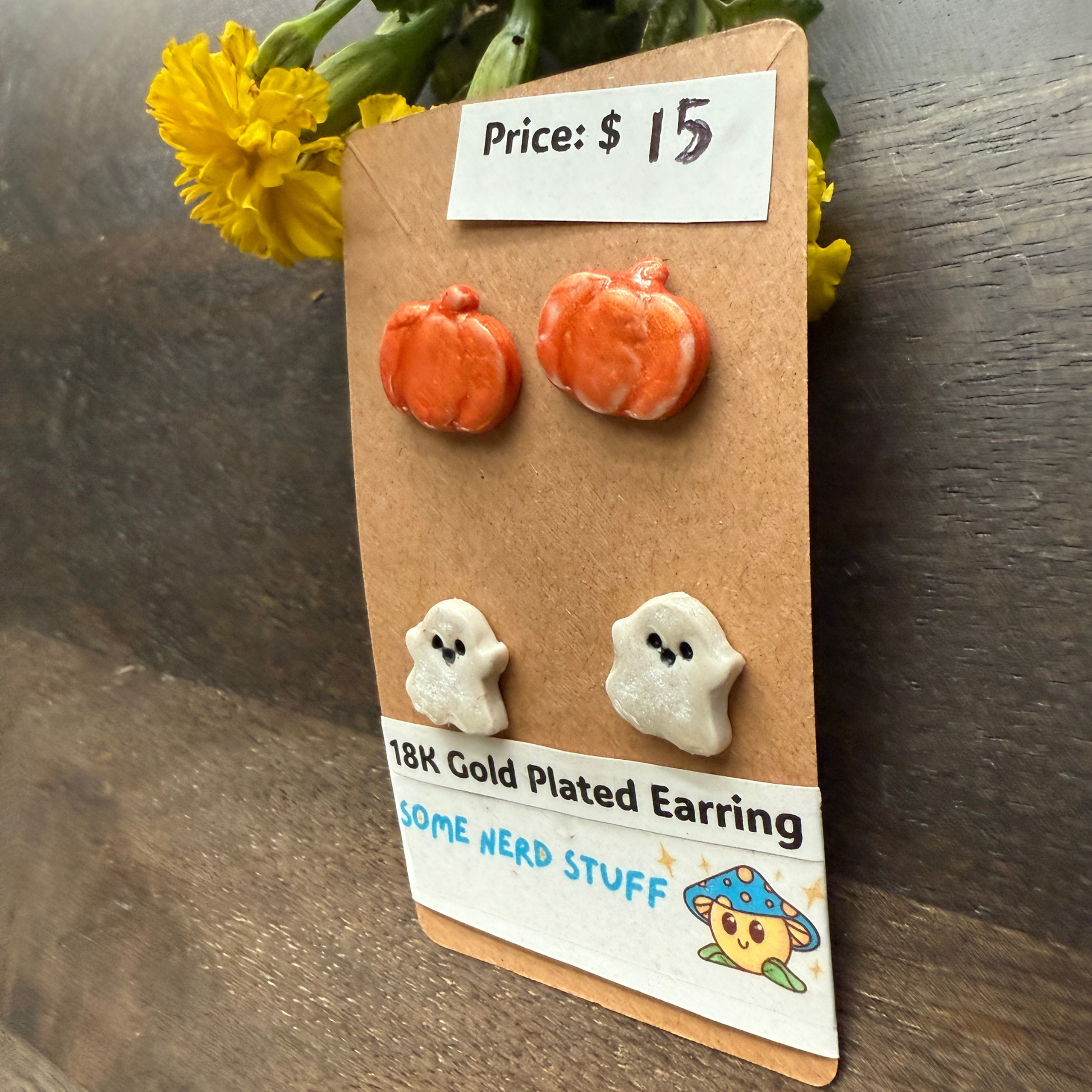 Halloween-themed earrings on a cardboard backing with a price tag, on a wooden surface featuring pumpkins and happy ghosts 18k gold plated earrings.