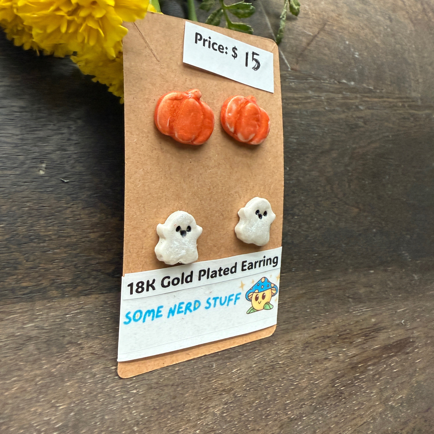 Halloween-themed earrings on a cardboard backing with a price tag, on a wooden surface featuring pumpkins and happy ghosts 18k gold plated earrings.