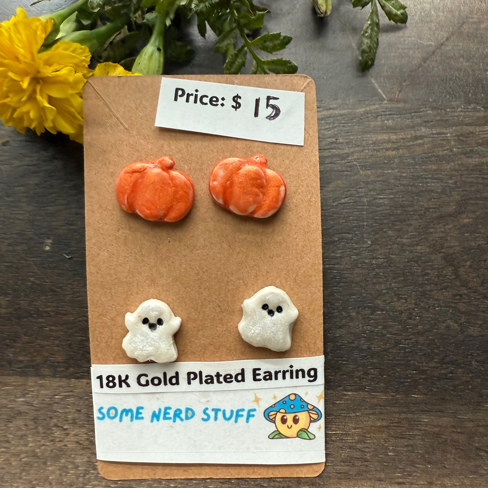 Halloween-themed earrings on a cardboard backing with a price tag, on a wooden surface featuring pumpkins and happy ghosts 18k gold plated earrings.