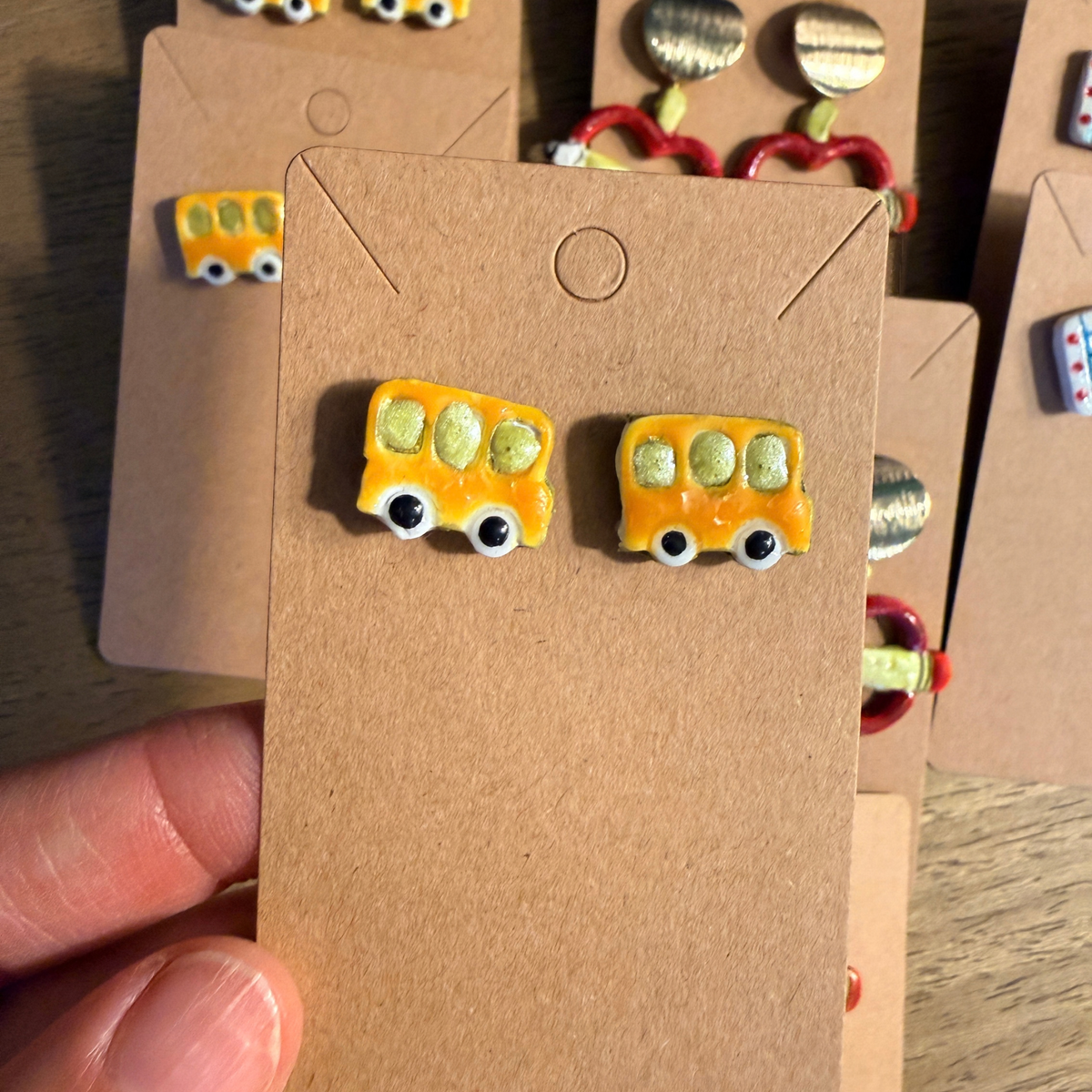 Hand holding a card with yellow school bus stud earrings featuring small windows and black wheels.