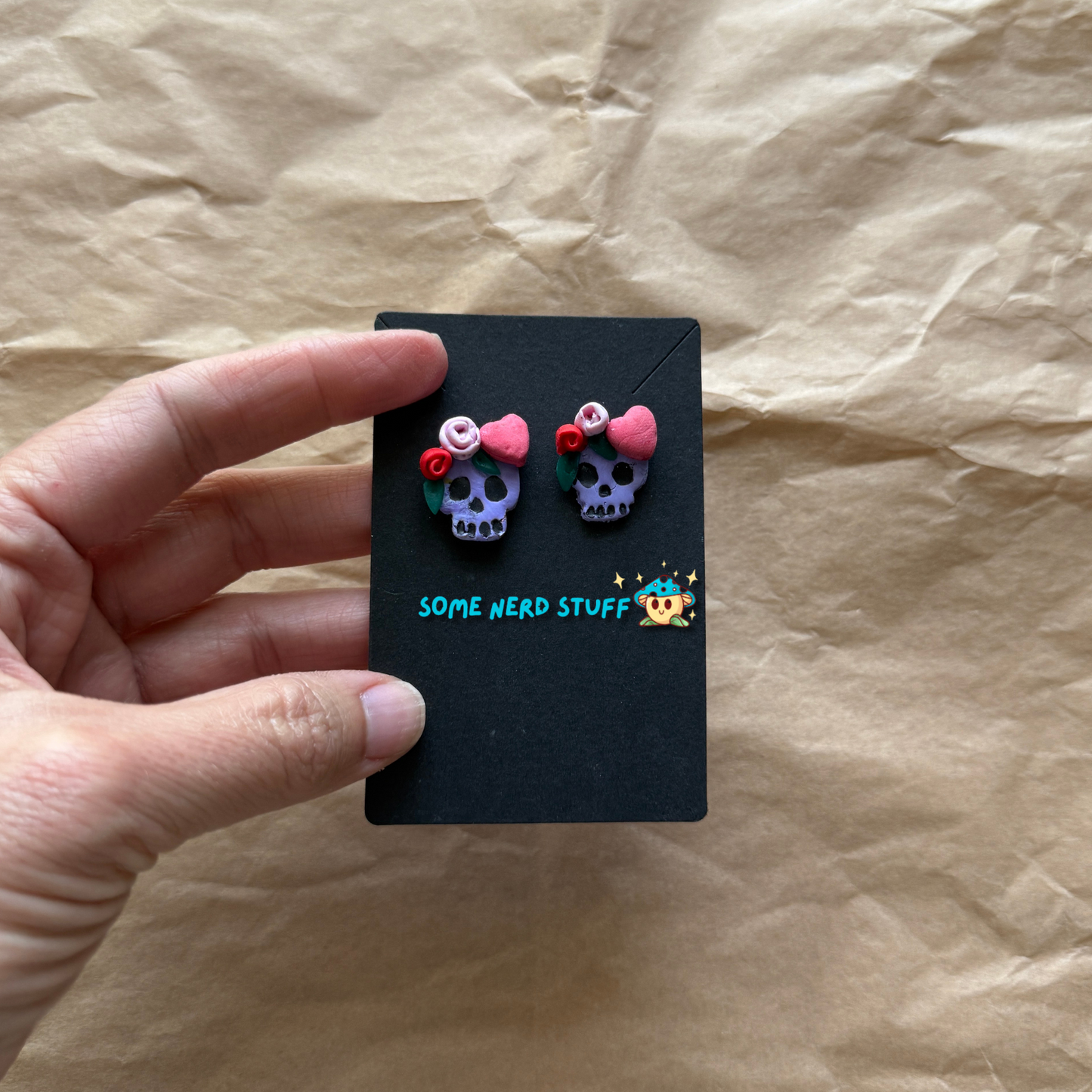 Polymer Clay Stud earrings with purple skulls, pink and red roses, with pink hearts hand holding to show size made by SomeNerdStuff