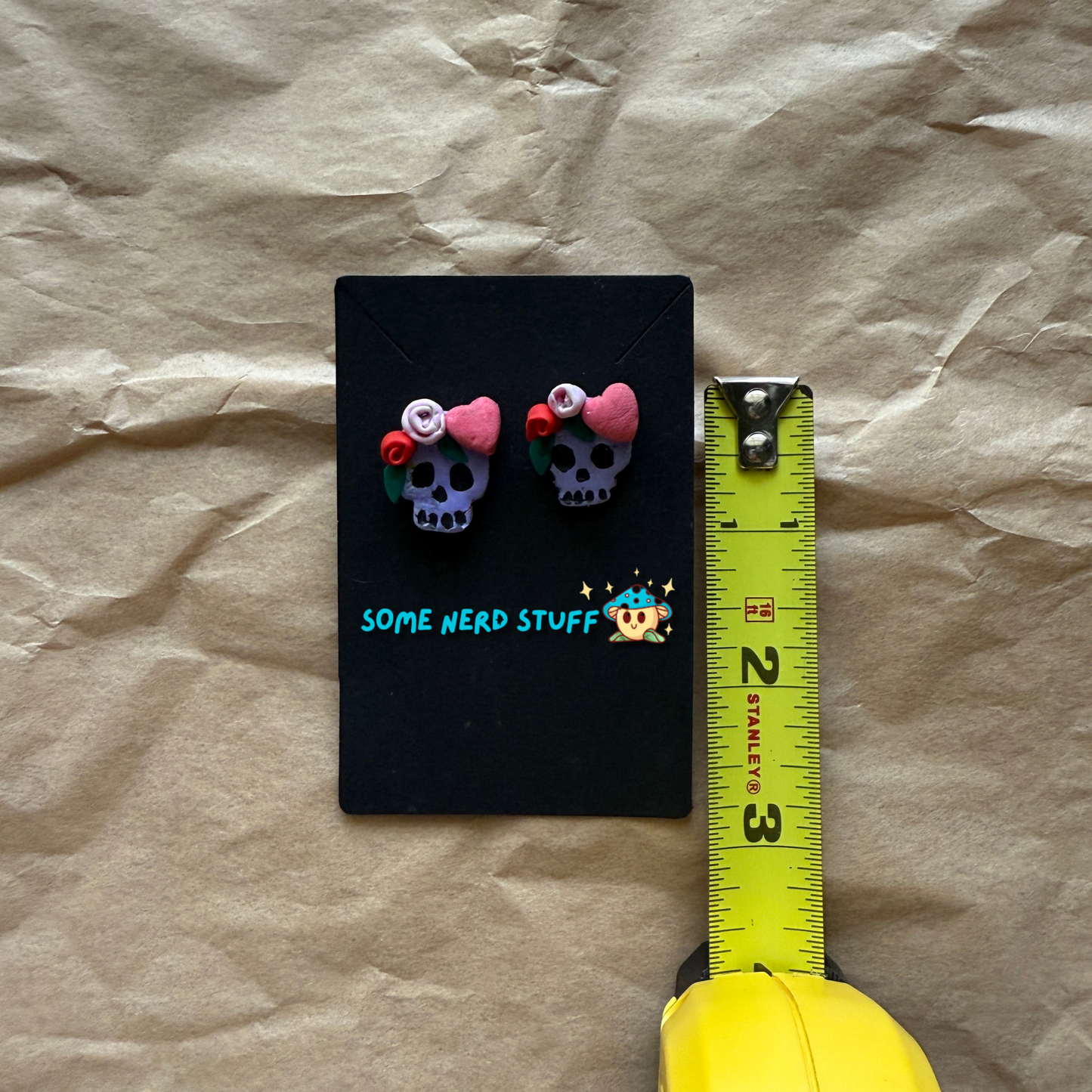 Polymer Clay Stud earrings with purple skulls, pink and red roses, with pink hearts shown with measuring tape made by SomeNerdStuff