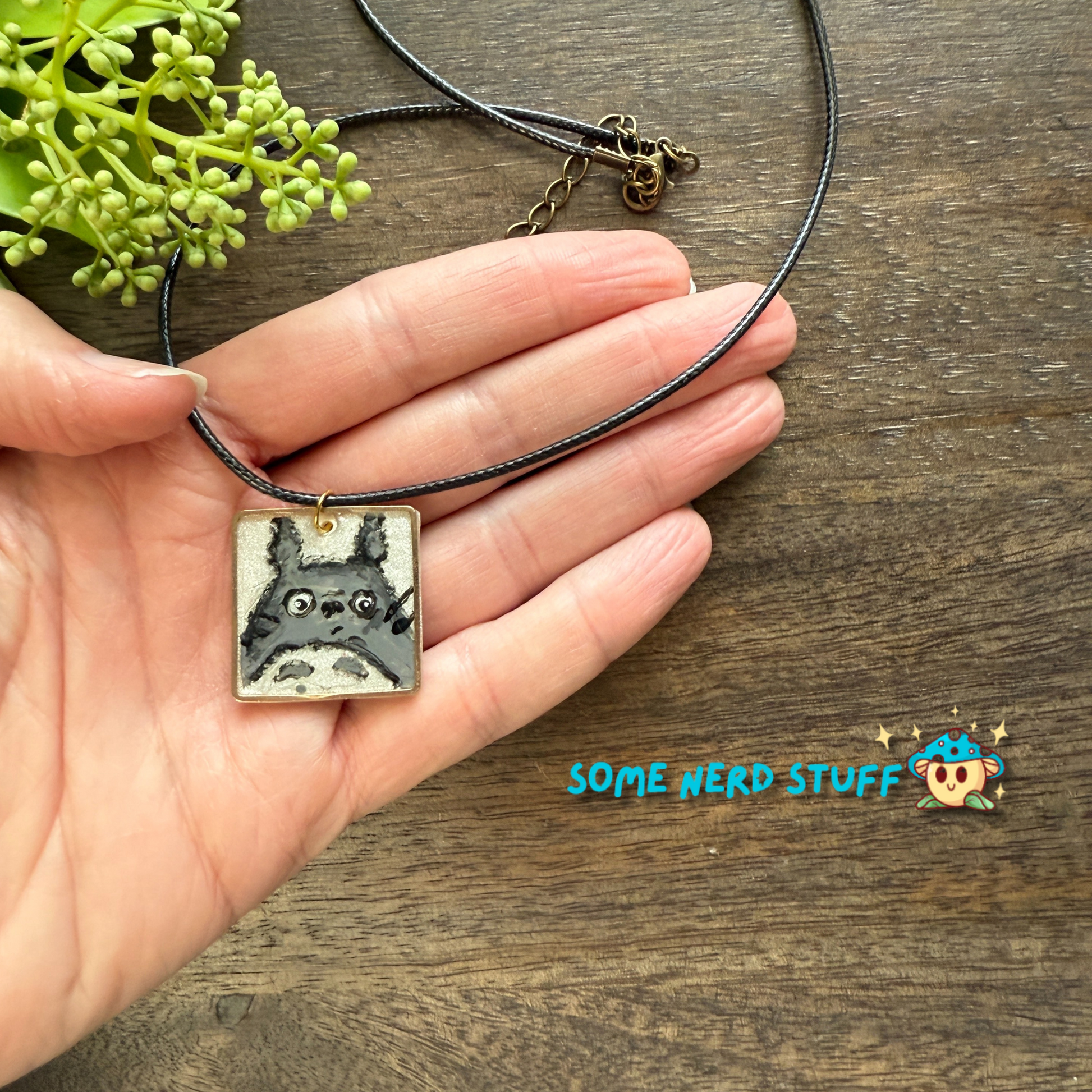 Acrylic hand painted Charm totoro in a metal square frame handing from a necklace with clasps shown in hand for size made by SomeNerdStuff