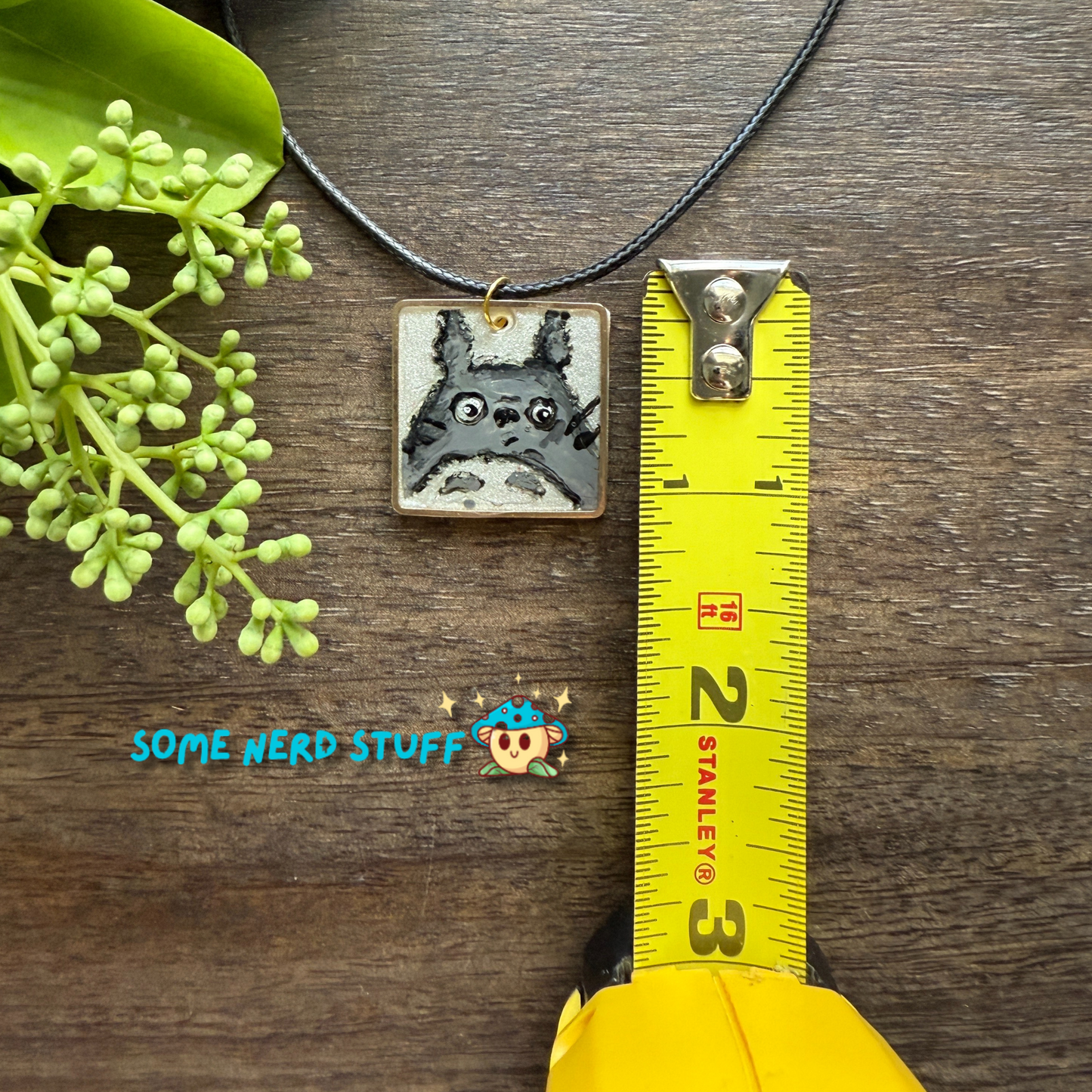 Acrylic hand painted Charm totoro in a metal square frame handing from a necklace with clasps shown with measuring tape made by SomeNerdStuff