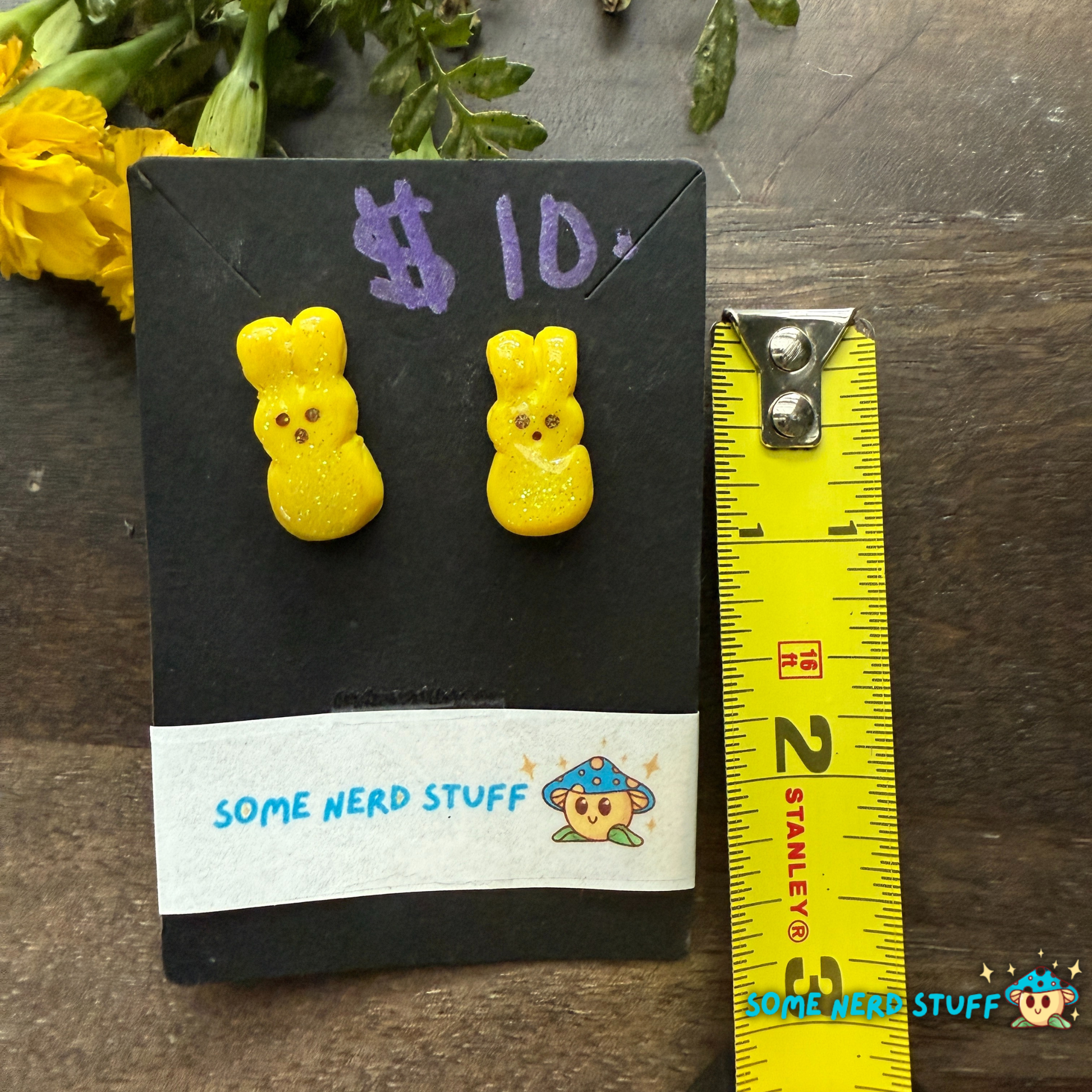 Yellow bunny-shaped earrings on a card with a measuring tape for scale, on a wooden surface.