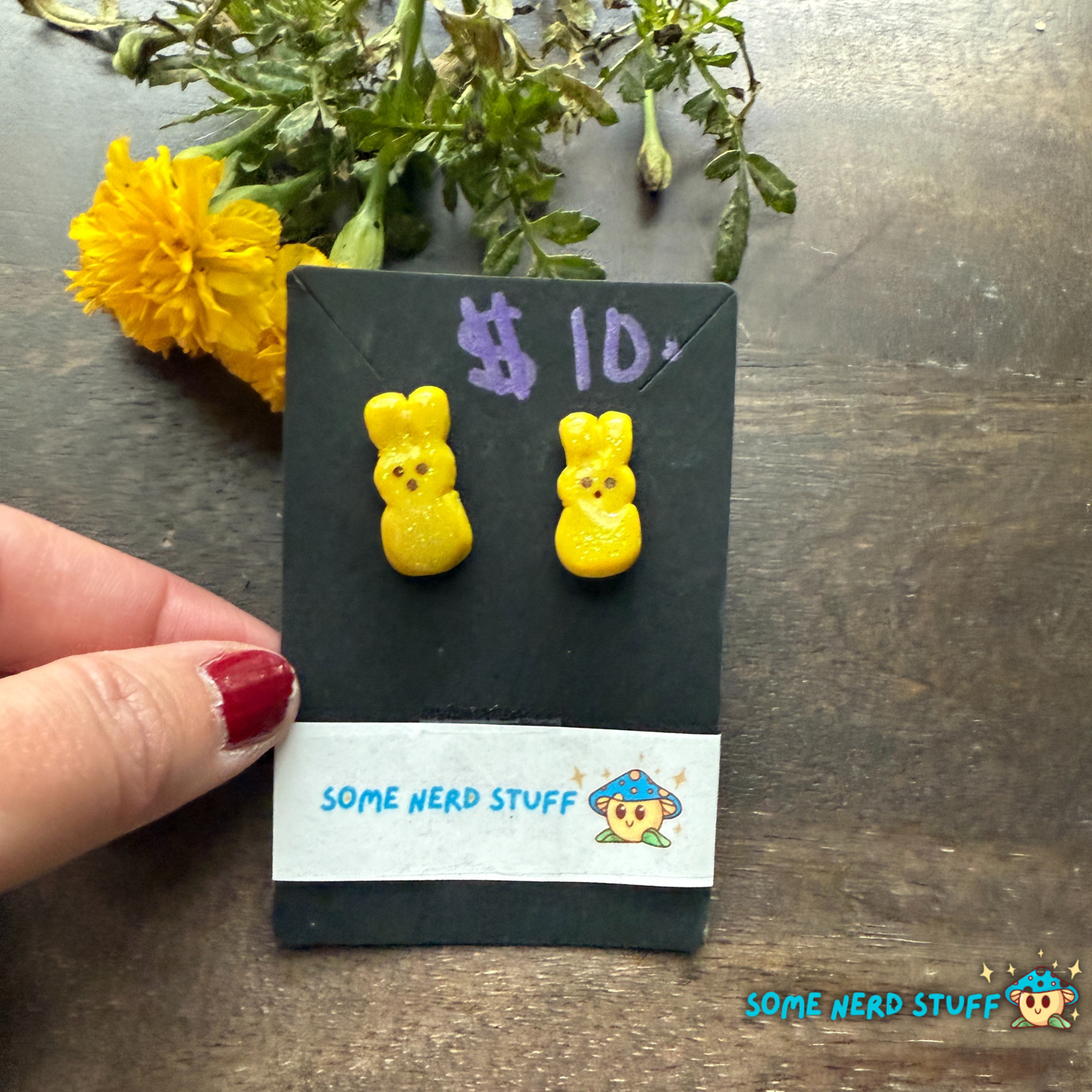 Yellow bunny-shaped earrings on a card labeled '$10' with 'Some Nerd Stuff' branding, against a dark background with yellow flowers.