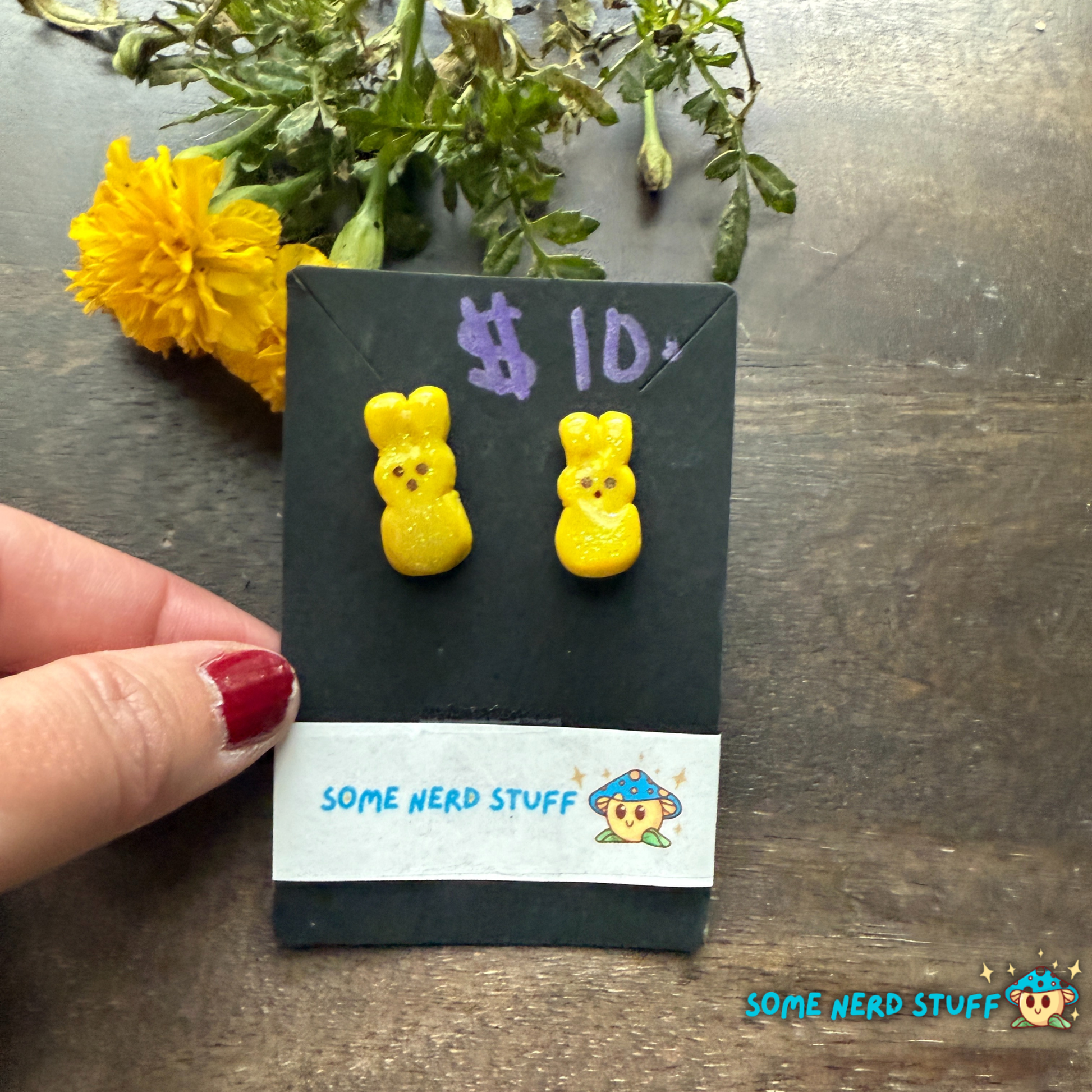 Yellow bunny-shaped earrings on a card labeled '$10' with 'Some Nerd Stuff' branding, against a dark background with yellow flowers.