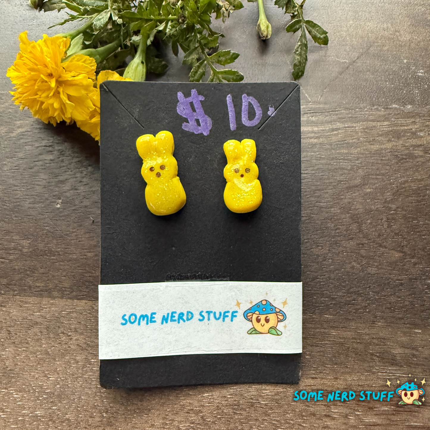 Yellow bear-shaped earrings on a card labeled 'Some Nerd Stuff' with a price tag, on a wooden surface with flowers.