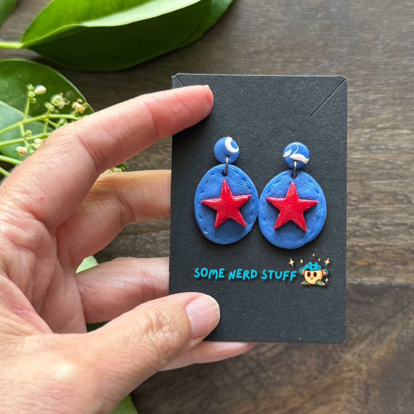 Super hero polymer clay inspired blue and red star earrings shown with hand to show size made by SomeNerdStuff