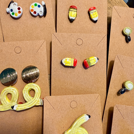 Several pairs of chunky baby pencil stud earrings displayed beside other school-themed designs.