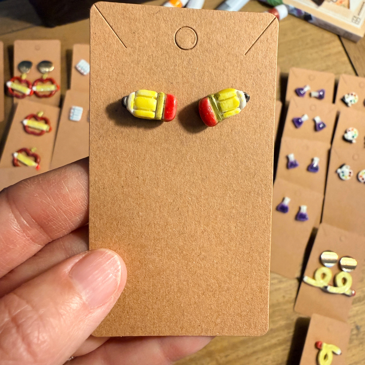 Hand holding a pair of short yellow pencil stud earrings with red eraser details.
