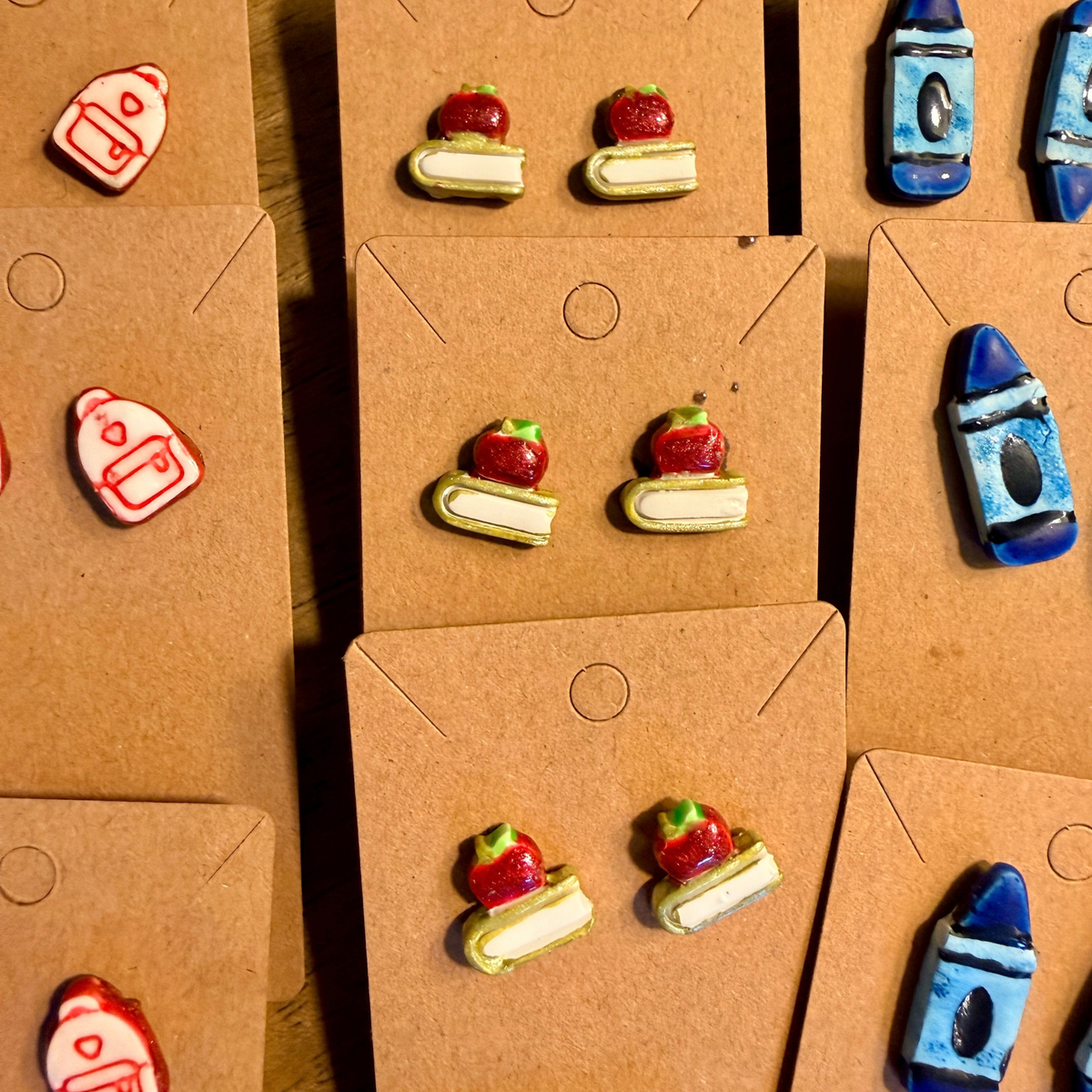Several pairs of apple and book stud earrings displayed beside backpack and blue crayon earrings.