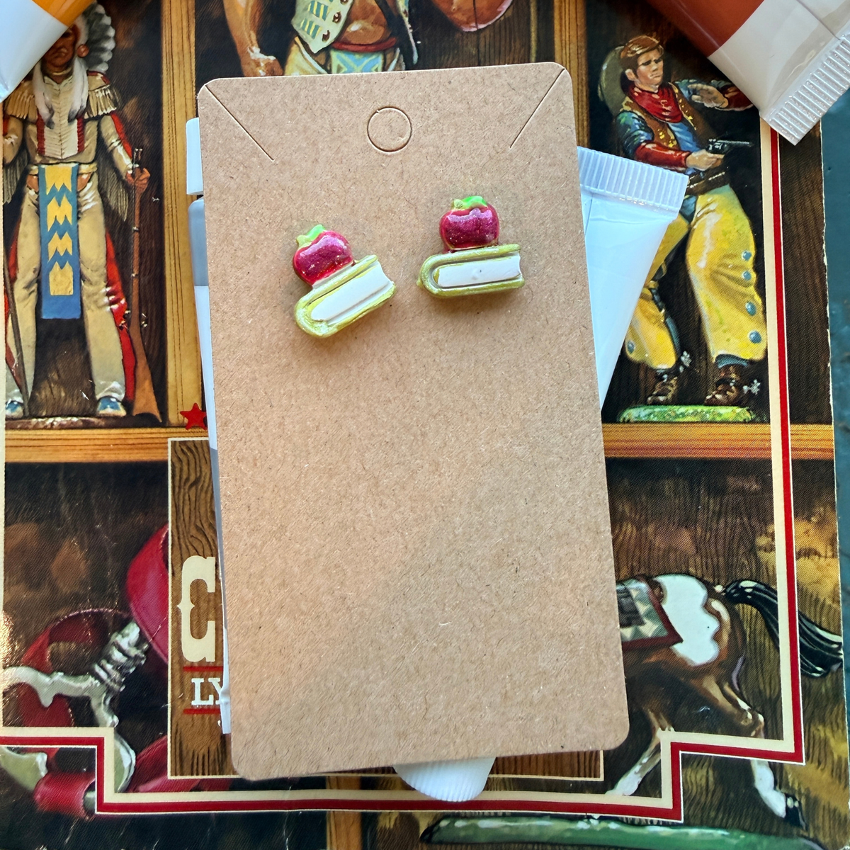 Pair of red apple and miniature book stud earrings displayed on a kraft card.